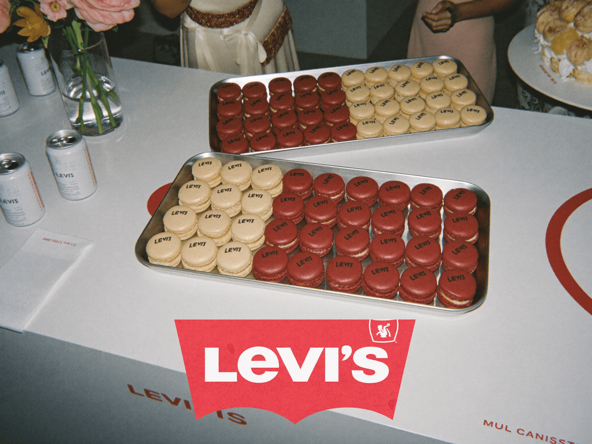 Dos charolas metálicas con macarons rojos y beige impresos con el logotipo de Levi’s, sobre una barra blanca rotulada con la marca, junto a latas y un arreglo floral.