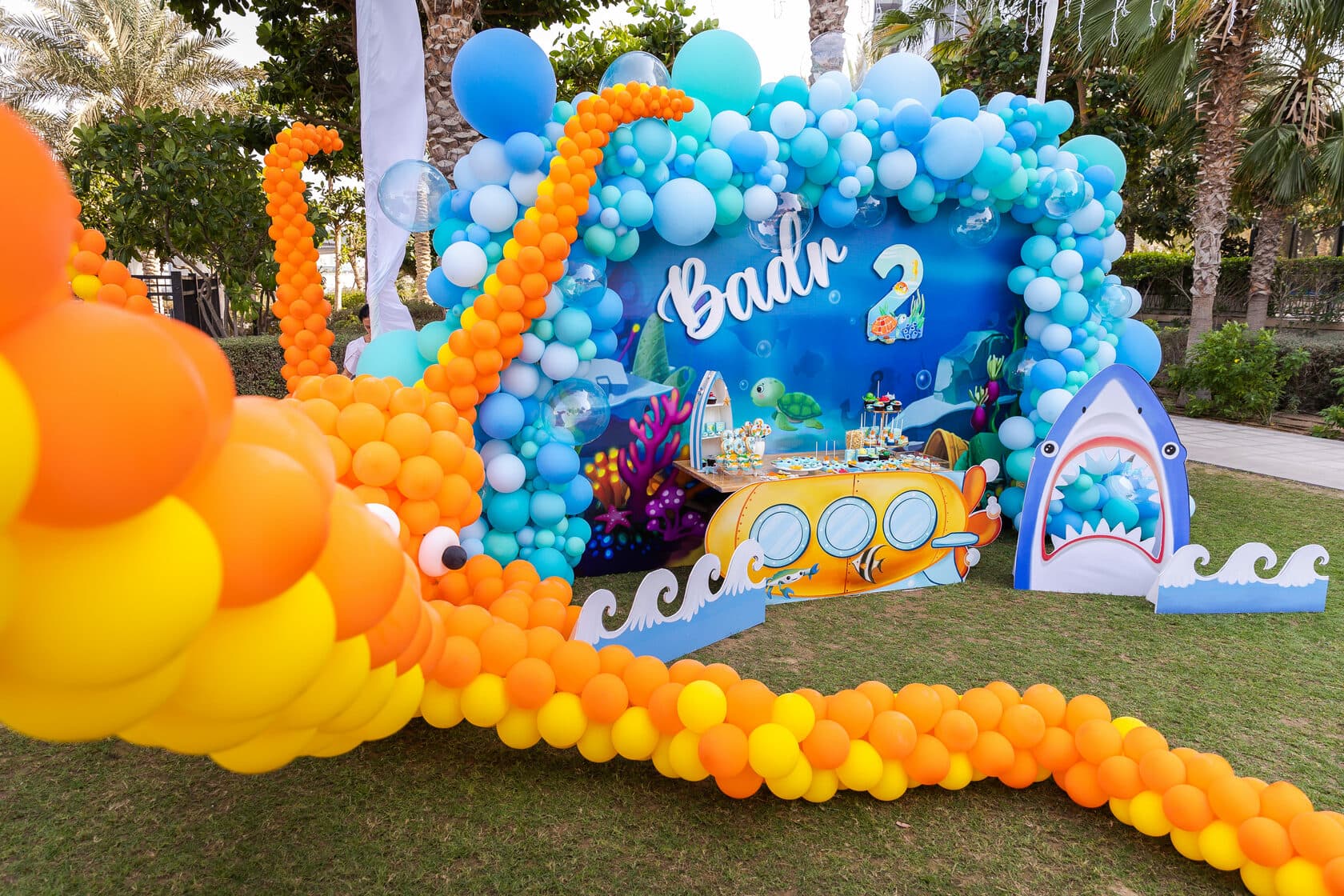 Arco de globos azules, naranjas y amarillos montado sobre el césped alrededor de un decorado de fondo marino con un submarino amarillo, un tiburón de cartón y el nombre «Badr» junto a un número dos.